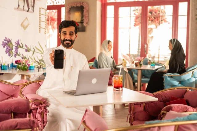 عرض خلفية سوداء على هاتف محمول في كوفي شوب راقي، قضاء وقت الفراغ باحتساء المشروب المفضل،  الاستمتاع بالمشروبات الباردة، صورة لرجل عربي خليجي سعودي يرتدي الثوب الأبيض يجلس في مقهى على طاولة حاسب محمول، إيماءات السعادة والسرور، تجلس في الخلفية نساء سعوديات