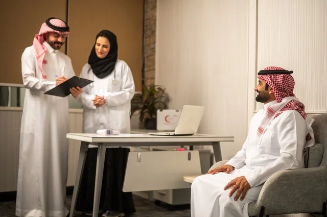 مساعد يقوم بتسجيل حالات المرضى, طبيبة عربية خليجيةة سعودية ترتدي المعطف الأبيض تقوم بشرح الحالة المرضية لمريض يرتدي الشماغ والثوب التقليدي، الحوار مع المرضى لكشف الحالة، تقديم الاستشارات في المراكز الطبيه, العمل في القطاعات الصحية، متابعة السجل الطبي للمراجعين