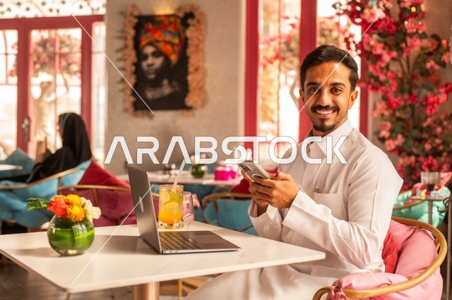 العمل على هاتف محمول في كوفي شوب راقي، قضاء وقت الفراغ باحتساء المشروب المفضل،  الاستمتاع بالمشروبات الباردة، صورة لرجل عربي خليجي سعودي يرتدي الثوب الأبيض يجلس في مقهى على طاولة حاسب محمول، إيماءات السعادة والسرور