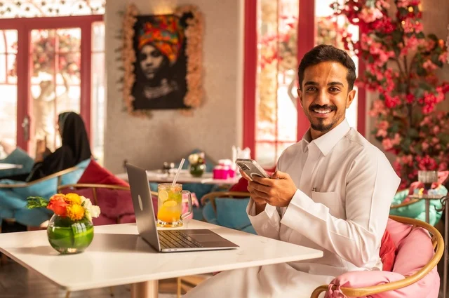 Working on a mobile phone in a classy coffee shop, spending free time sipping a favorite drink, enjoying cold beverages, a picture of a Saudi Arabian Gulf man wearing a white thobe sitting in a café at a laptop table, gestures of happiness and joy.