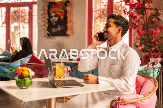 إجراء مكالمة هاتفية في كوفي شوب راقي، قضاء وقت الفراغ باحتساء المشروب المفضل،  الاستمتاع بالمشروبات الباردة، صورة لرجل عربي خليجي سعودي يرتدي الثوب الأبيض يجلس في مقهى على طاولة حاسب محمول، إيماءات السعادة والسرور