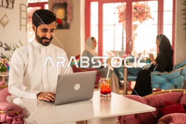 دمج التكنولوجيا مع الحياة اليومية  في كوفي شوب راقي، قضاء وقت الفراغ باحتساء المشروب المفضل،  الاستمتاع بالمشروبات الباردة، صورة لرجل عربي خليجي سعودي يرتدي الثوب الأبيض يجلس في مقهى على طاولة حاسب محمول، إيماءات السعادة والسرور، تجلس في الخلفية نساء سعوديات