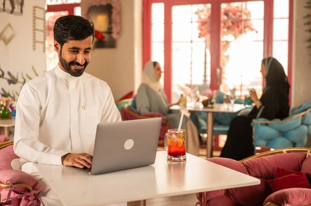 دمج التكنولوجيا مع الحياة اليومية  في كوفي شوب راقي، قضاء وقت الفراغ باحتساء المشروب المفضل،  الاستمتاع بالمشروبات الباردة، صورة لرجل عربي خليجي سعودي يرتدي الثوب الأبيض يجلس في مقهى على طاولة حاسب محمول، إيماءات السعادة والسرور، تجلس في الخلفية نساء سعوديات