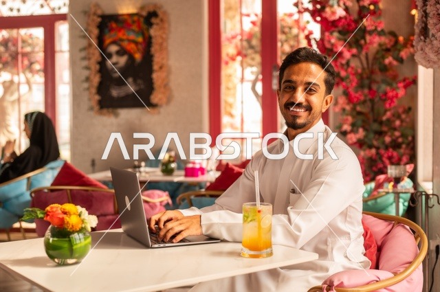 النظر إلى الكاميرا في كوفي شوب راقي، قضاء وقت الفراغ باحتساء المشروب المفضل،  الاستمتاع بالمشروبات الباردة، صورة لرجل عربي خليجي سعودي يرتدي الثوب الأبيض يجلس في مقهى على طاولة حاسب محمول، إيماءات السعادة والسرور