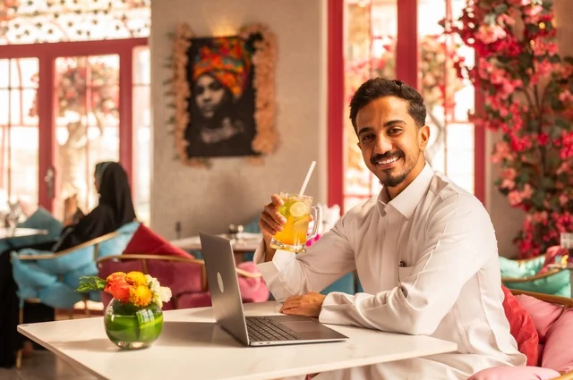 الاستمتاع بالمشروبات الباردة في كوفي شوب راقي، قضاء وقت الفراغ باحتساء المشروب المفضل،العمل عن بعد ، صورة لرجل عربي خليجي سعودي يرتدي الثوب الأبيض يجلس في مقهى على طاولة حاسب محمول، إيماءات السعادة والسرور