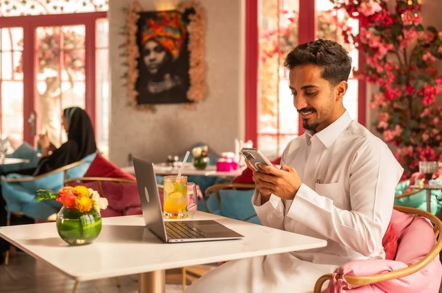 Working on a mobile phone in a classy coffee shop, spending free time sipping a favorite drink, enjoying cold beverages, a picture of a Saudi Arabian Gulf man wearing a white thobe sitting in a café at a laptop table, gestures of happiness and joy.