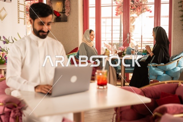 دمج التكنولوجيا مع الحياة اليومية  في كوفي شوب راقي، قضاء وقت الفراغ باحتساء المشروب المفضل،  الاستمتاع بالمشروبات الباردة، صورة لرجل عربي خليجي سعودي يرتدي الثوب الأبيض يجلس في مقهى على طاولة حاسب محمول، إيماءات السعادة والسرور، تجلس في الخلفية نساء سعوديات