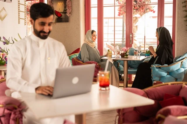 Integrating technology with daily life in a classy coffee shop, spending leisure time sipping a favorite drink, enjoying cold beverages, a picture of a Saudi Arabian Gulf man wearing a white thobe sitting in a café at a laptop table, gestures of happiness and joy, with Saudi women sitting in the background.