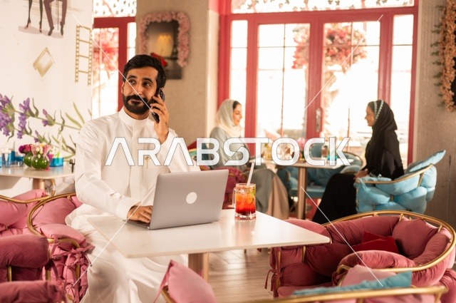 إجراء مكالمة هاتفية في كوفي شوب راقي، قضاء وقت الفراغ باحتساء المشروب المفضل،  الاستمتاع بالمشروبات الباردة، صورة لرجل عربي خليجي سعودي يرتدي الثوب الأبيض يجلس في مقهى على طاولة حاسب محمول، إيماءات السعادة والسرور