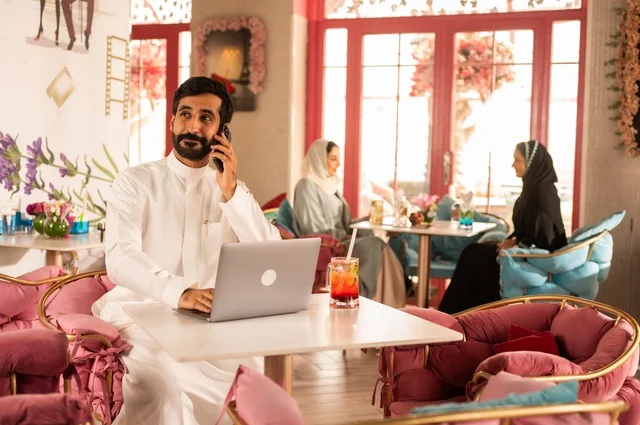 إجراء مكالمة هاتفية في كوفي شوب راقي، قضاء وقت الفراغ باحتساء المشروب المفضل،  الاستمتاع بالمشروبات الباردة، صورة لرجل عربي خليجي سعودي يرتدي الثوب الأبيض يجلس في مقهى على طاولة حاسب محمول، إيماءات السعادة والسرور