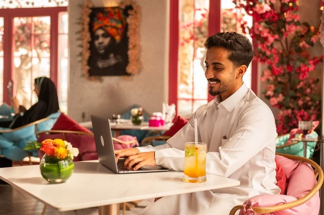 Working remotely in an upscale coffee shop, spending free time sipping a favorite drink, enjoying cold beverages, a picture of a Saudi Arabian Gulf man wearing a white thobe sitting in a café at a laptop table, gestures of happiness and joy.