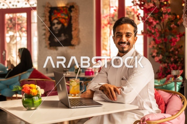 Looking at the camera in a fancy coffee shop, spending free time sipping a favorite drink, enjoying cold beverages, a picture of a Saudi Arabian Gulf man wearing a white thobe sitting in a café at a laptop table, gestures of happiness and joy.