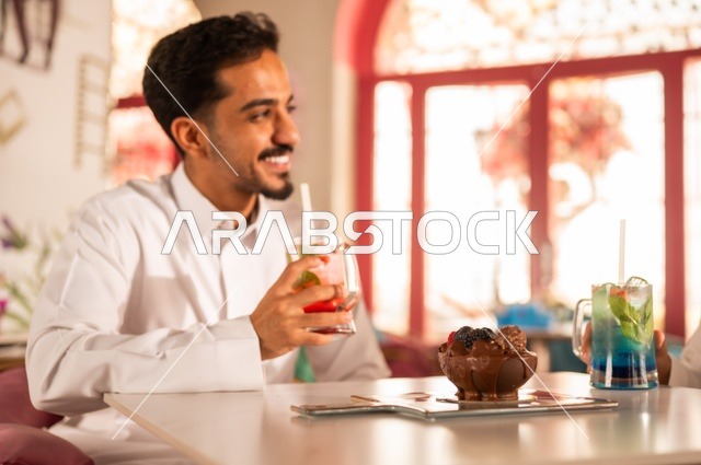 صورة مقربة جانبية لشاب  في كوفي شوب راقي، قضاء وقت الفراغ باحتساء المشروب المفضل،  الاستمتاع بالمشروبات الباردة، رجل عربي خليجي سعودي يرتدي الثوب الأبيض يجلس في مقهى على طاولة ، إيماءات السعادة والسرور