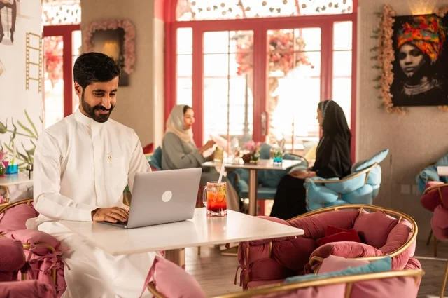 استخدام التكنولوجيا في المهام اليومية في كوفي شوب راقي، قضاء وقت الفراغ باحتساء المشروب المفضل،  الاستمتاع بالمشروبات الباردة، صورة لرجل عربي خليجي سعودي يرتدي الثوب الأبيض يجلس في مقهى على طاولة حاسب محمول، إيماءات السعادة والسرور، تجلس في الخلفية نساء سعوديات