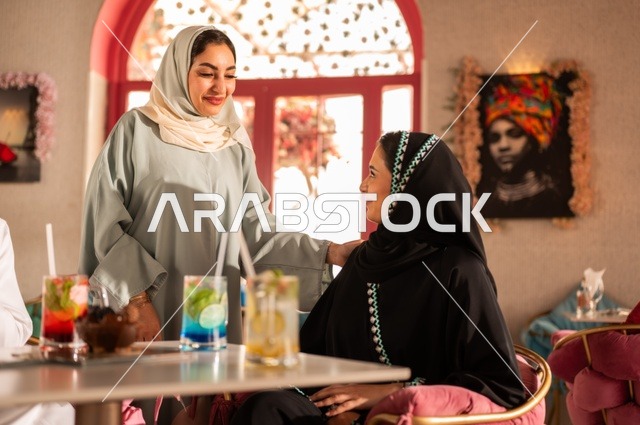 تعابير وجه ويد بإيماءات السعادة و السرور, التنزه مع الأصدقاء، قضاء الأوقات الممتعة في الخارج، الاستمتاع بالأنشطة الترفيهية, الاستمتاع بالمشروبات الباردة في فصل الصيف,  صديقتان عربياتان خليجيتان سعوديتان يرتدون عباءة وحجاب داخل كافي شوب راقي