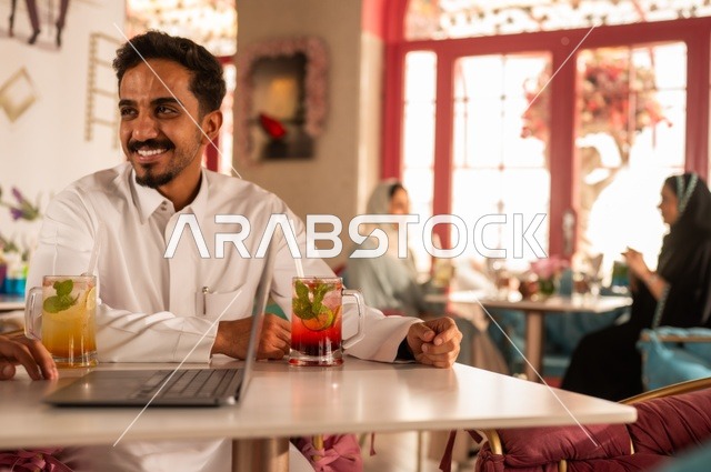 قضاء الأوقات الممتعة في الخارج، الاستمتاع بالأنشطة الترفيهية, الاستمتاع بالمشروبات الباردة في فصل الصيف,  شاب عربي خليجي سعودي يرتدي الثوب التقليدي يجلس في كافي شوب راقي ،التنزه مع الأصدقاء، إيماءات السعادة و السرور, حاسب محمول على الطاولة،خلفية نساء في مقهى .
