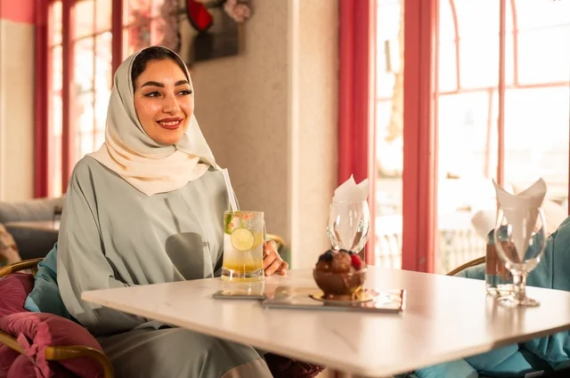 الاستمتاع بالأنشطة الترفيهية, الاستمتاع بالمشروبات الباردة في فصل الصيف,  امرأة عربية خليجية سعودية محجبة ترتدي عباءة تجلس في كافي شوب راقي ،التنزه مع الأصدقاء، إيماءات السعادة و السرور, قضاء الأوقات الممتعة في الخارج .