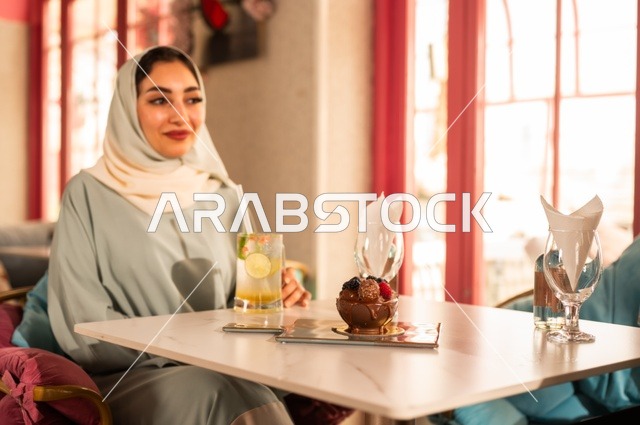 الاستمتاع بالأنشطة الترفيهية, الاستمتاع بالمشروبات الباردة في فصل الصيف,  امرأة عربية خليجية سعودية محجبة ترتدي عباءة تجلس في كافي شوب راقي ،التنزه مع الأصدقاء، إيماءات السعادة و السرور, قضاء الأوقات الممتعة في الخارج .