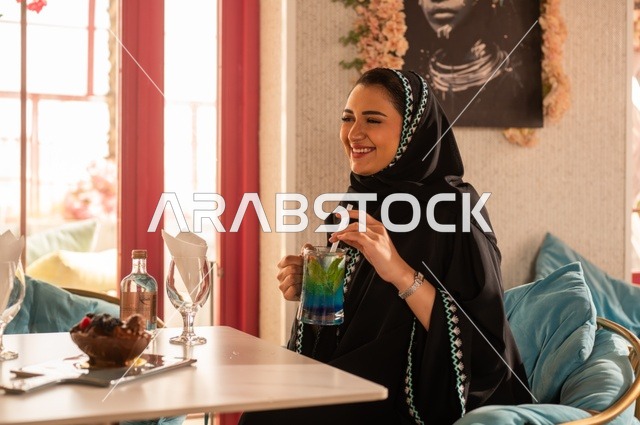 Enjoying cold drinks in the summer, enjoying recreational activities, a Saudi Gulf Arab woman wearing a hijab and an abaya sitting in a classy café, hanging out with friends, gestures of happiness and joy, spending enjoyable times outdoors.