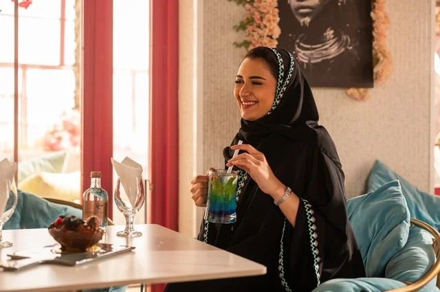 Enjoying cold drinks in the summer, enjoying recreational activities, a Saudi Gulf Arab woman wearing a hijab and an abaya sitting in a classy café, hanging out with friends, gestures of happiness and joy, spending enjoyable times outdoors.
