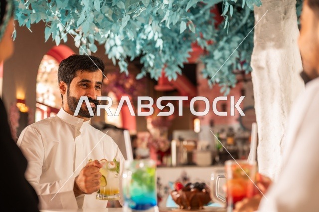 Discussion and dialogue in a calm atmosphere, spending enjoyable times with friends, sharing cold drinks and sweets, building successful social relationships among people, a close-up image of two smiling Saudi Gulf Arab colleagues sitting in one of the upscale cafes with expressions of happiness and joy.