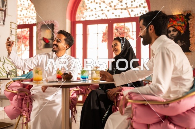 Selfie of beautiful moments, building successful social relationships between people, discussion and dialogue in calm atmospheres, spending enjoyable times with friends, sharing cold drinks and sweets, a picture of two smiling Saudi Gulf Arab colleagues sitting in one of the upscale cafes with expressions of happiness and joy.