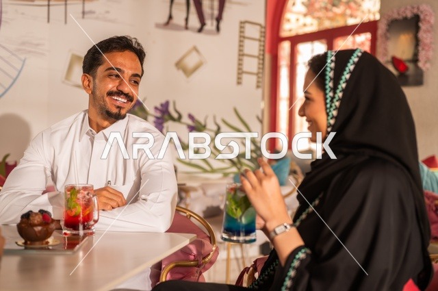 مفهوم المودة والالفة ، ايماءات السرور والسعادة ، الاستمتاع بشرب المشروبات الباردة ، قضاء أوقات ممتعة ورومانسية، زوجان عربيان خليجيان سعوديان مبتسمان يجلسان في المقهى