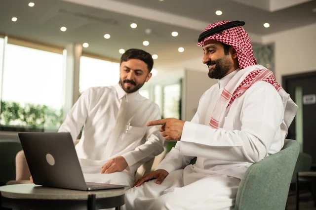 تبادل الخبرات والافكار، دراسة وتحليل المشاريع الجديدة، استخدام جهاز تقني حديث، الوحدة والعمل الجماعي، تطوير العمل وحل المشكلات، رجل عربي خليجي سعودي يرتدي الشماغ والثوب التقليدي يقوم بالعمل برفقة زميله في الخارج، مهنة ووظيفة سعودية مكتبية
