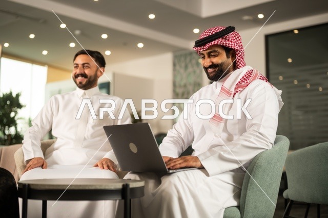 تبادل الخبرات والافكار، دراسة وتحليل المشاريع الجديدة، استخدام جهاز تقني حديث، الوحدة والعمل الجماعي، تطوير العمل وحل المشكلات، رجل عربي خليجي سعودي يرتدي الشماغ والثوب التقليدي يقوم بالعمل برفقة زميله في الخارج، مهنة ووظيفة سعودية مكتبية