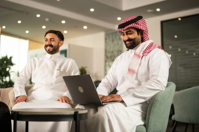 Exchange of experiences and ideas, studying and analyzing new projects, using modern technical devices, unity and teamwork, developing work and solving problems, an Arab Gulf Saudi man wearing the traditional headscarf and thobe working alongside his colleague abroad, a Saudi office profession and job.