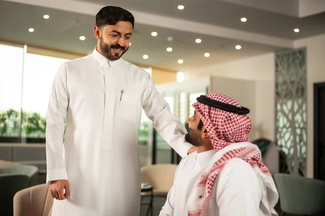 Exchange of experiences and ideas, studying and analyzing new projects, using modern technical devices, unity and teamwork, developing work and solving problems, an Arab Gulf Saudi man wearing the traditional headscarf and thobe working alongside his colleague abroad, a Saudi office profession and job.