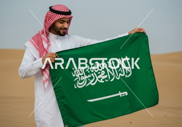 الفخر والاعتزاز بالهوية الوطنية، رجل عربي خليجي سعودي يرتدي الثوب التقليدي والشماغ يقف في الصحراء يحمل علم المملكة العربية السعودية، ذكرى العيد الوطني السعودي 23 مارس، يوم العلم السعودي 11 مارس، حب الوطن والانتماء إليه