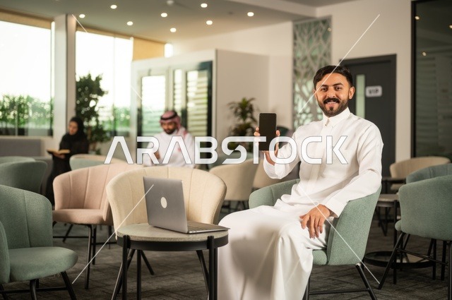 عرض شاشة هاتف محمول سوداء، إدارة شؤون الأعمال وتنظيمها عبر جهاز تقني حديث، إنجاز مهام العمل عن بعد، رجل عربي خليجي سعودي يرتدي الثوب التقليدي يجلس  في إحدى المقاهي الراقية ويستخدم الحاسوب المحمول