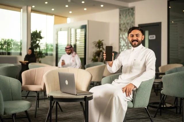 A black mobile phone screen, managing and organizing business affairs through a modern technical device, completing work tasks remotely, an Arab Gulf Saudi man wearing traditional attire sitting in a high-end café using a laptop.