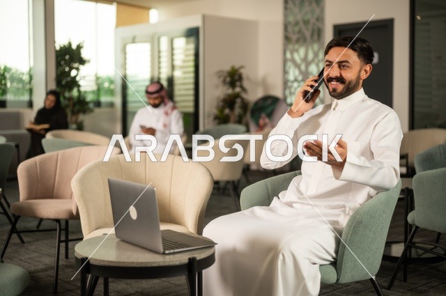اجراء مكالمة هاتفية عبر الهاتف المحمول، إدارة شؤون الأعمال وتنظيمها عبر جهاز تقني حديث، إنجاز مهام العمل عن بعد، رجل عربي خليجي سعودي يرتدي الثوب التقليدي يجلس  في إحدى المقاهي الراقية ويستخدم الحاسوب المحمول