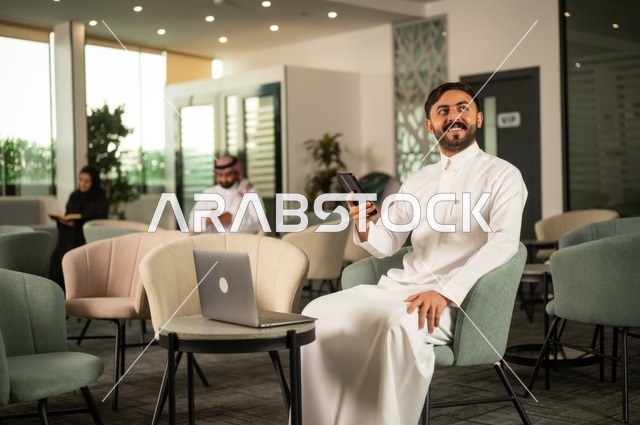 Working on a mobile phone, managing and organizing business affairs through a modern technological device, completing remote work tasks, an Arab Gulf Saudi man wearing traditional attire sitting in a high-end café using a laptop.