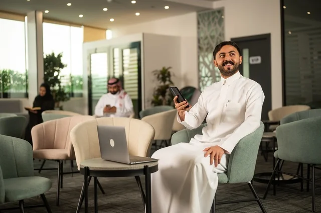 Working on a mobile phone, managing and organizing business affairs through a modern technological device, completing remote work tasks, an Arab Gulf Saudi man wearing traditional attire sitting in a high-end café using a laptop.