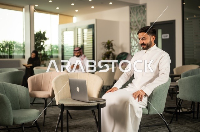 إدارة شؤون الأعمال وتنظيمها عبر جهاز تقني حديث، إنجاز مهام العمل عن بعد، رجل عربي خليجي سعودي يرتدي الثوب التقليدي يجلس  في إحدى المقاهي الراقية ويستخدم الحاسوب المحمول
