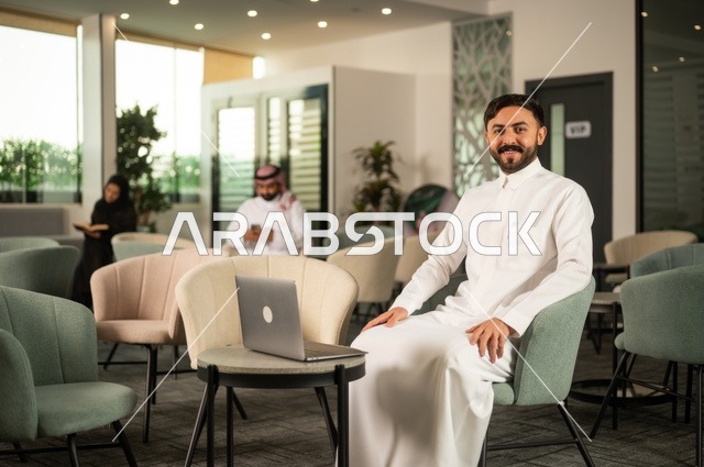 إدارة شؤون الأعمال وتنظيمها عبر جهاز تقني حديث، إنجاز مهام العمل عن بعد، رجل عربي خليجي سعودي يرتدي الثوب التقليدي يجلس  في إحدى المقاهي الراقية ويستخدم الحاسوب المحمول