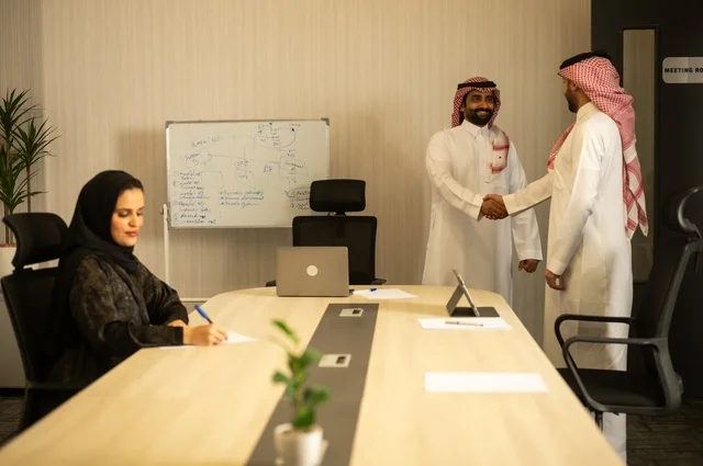 عقد صفقات عمل جديدة ناجحة بين رجال الأعمال، صداقات وتعارف وبناء علاقات اجتماعية، الشراكة في الأعمال والمشاريع، رجلان عربيان خليجيان سعوديان يرتديان الشماغ والثوب التقليدي يقومان بمصافحة بعضهما، توسيع مجالات العمل، إمرأة ترتدي الحجاب والعباءة تدون البيانات على ورقة