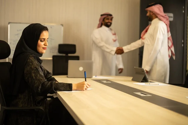 عقد صفقات عمل جديدة ناجحة بين رجال الأعمال، صداقات وتعارف وبناء علاقات اجتماعية، الشراكة في الأعمال والمشاريع، رجلان عربيان خليجيان سعوديان يرتديان الشماغ والثوب التقليدي يقومان بمصافحة بعضهما، توسيع مجالات العمل، إمرأة ترتدي الحجاب والعباءة تدون البيانات على ورقة