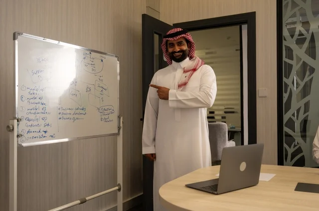 Ensuring a clear and comprehensive understanding of the business, a Saudi Arabian Gulf Arab man wearing a ghutrah and traditional thobe is using a whiteboard to explain the processes and procedures followed in the company, teamwork and team spirit, discussing work plans, using a laptop to take notes, professionalism, and dedication to achievement.