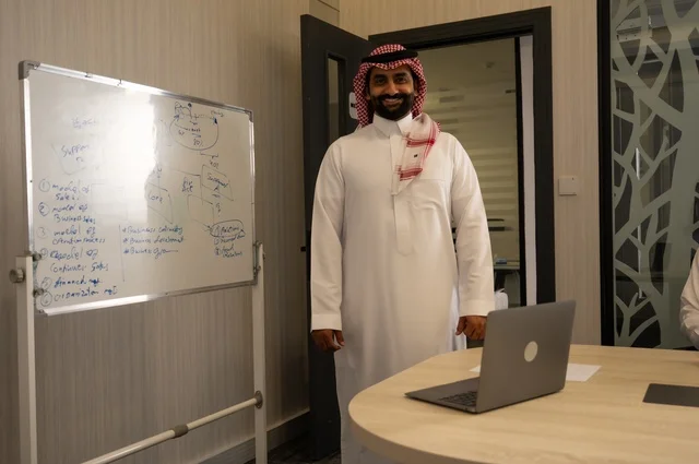 ضمان فهم واضح وشامل للاعمال، رجل عربي خليجي سعودي يرتدي الشماغ والثوب التقليدي يقوم باستعمال السبورة لتوضيح العمليات والاجراءات المتبعة في الشركة، العمل الجماعي وروح الفريق، مناقشة خطط العمل، استخدام اللابتوب في تدوين الملاحظات، الاحترافية والتفاني في الإنجاز