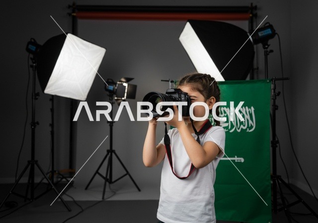 استخدام كاميرات رقمية عالية الدقة, الطموح في تحقيق الآمال والأمنيات، ممارسة هواية التصوير, طفلة عربية خليجية سعودية ترتدي كنزة بيضاء تمسك بيدها كاميرا فوتوغرافية, مصور محترف يقوم بالتقاط صور تذكارية, في الخلفية استوديو تصوير وعلم المملكة العربية السعودية