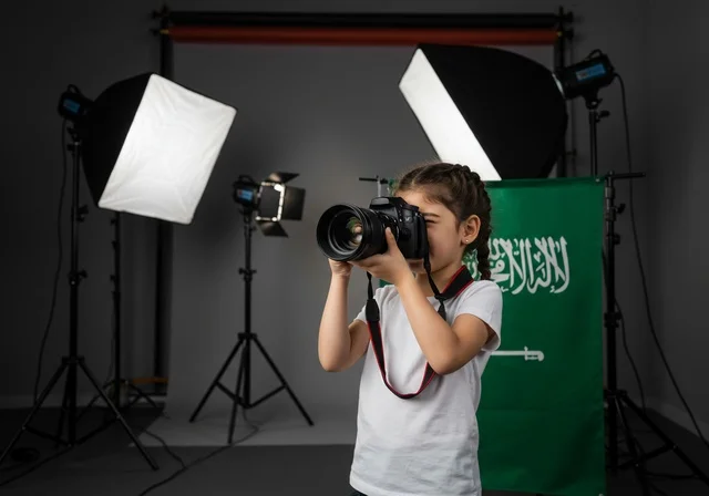 استخدام كاميرات رقمية عالية الدقة, الطموح في تحقيق الآمال والأمنيات، ممارسة هواية التصوير, طفلة عربية خليجية سعودية ترتدي كنزة بيضاء تمسك بيدها كاميرا فوتوغرافية, مصور محترف يقوم بالتقاط صور تذكارية, في الخلفية استوديو تصوير وعلم المملكة العربية السعودية