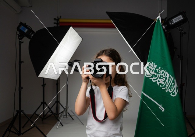 استخدام كاميرات رقمية عالية الدقة, الطموح في تحقيق الآمال والأمنيات، ممارسة هواية التصوير, طفلة عربية خليجية سعودية ترتدي كنزة بيضاء تمسك بيدها كاميرا فوتوغرافية, مصور محترف يقوم بالتقاط صور تذكارية, في الخلفية استوديو تصوير وعلم المملكة العربية السعودية