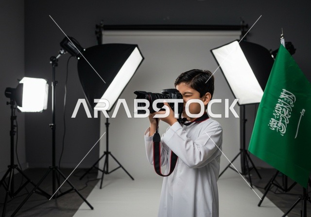 ممارسة هواية التصوير, استخدام كاميرات رقمية عالية الدقة, الطموح في تحقيق الآمال والأمنيات، طفل عربي خليجي سعودي يرتدي ثوب أبيض يمسك بيده كاميرا فوتوغرافية, مصور محترف يقوم بالتقاط صور تذكارية, في الخلفية استوديو تصوير وعلم المملكة العربية السعودية