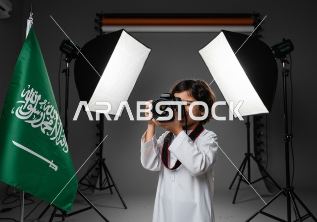 ممارسة هواية التصوير, استخدام كاميرات رقمية عالية الدقة, الطموح في تحقيق الآمال والأمنيات، طفل عربي خليجي سعودي يرتدي ثوب أبيض يمسك بيده كاميرا فوتوغرافية, مصور محترف يقوم بالتقاط صور تذكارية, في الخلفية استوديو تصوير وعلم المملكة العربية السعودية