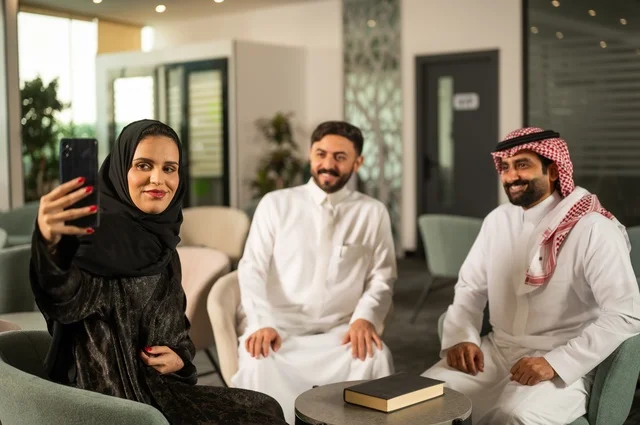 Friendly introductory sessions, colleagues at a meeting outside the company, an Arab Gulf Saudi woman wearing a hijab and abaya using her mobile phone with her friends, taking commemorative selfies via mobile phone, documenting happy moments.