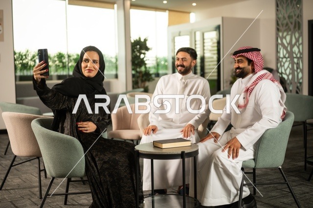Friendly introductory sessions, colleagues at a meeting outside the company, an Arab Gulf Saudi woman wearing a hijab and abaya using her mobile phone with her friends, taking commemorative selfies via mobile phone, documenting happy moments.